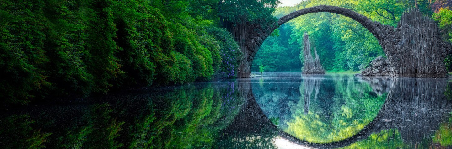 Arch bridge reflected over a calm river with green foliage surrounding